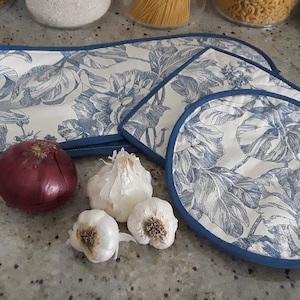 Puede incluir: Conjunto de cuatro manoplas para horno con estampado floral azul y blanco. Las manoplas están hechas de tela acolchada con ribete azul. Las manoplas están dispuestas sobre una encimera gris claro con una cebolla roja y cuatro dientes de ajo.