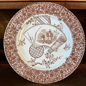 May include: A white ceramic plate with a brown floral pattern border. The center of the plate features a brown and white design with a fan, a scroll, and a camel caravan.