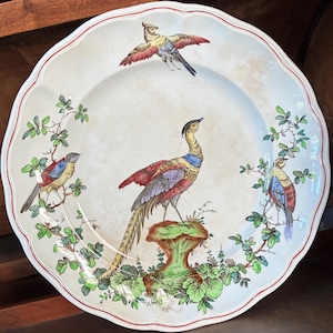 May include: A round ceramic plate with a scalloped edge and a thin red rim. The center features a large bird perched on a green pedestal, surrounded by two smaller birds and green leafy branches. The background is off-white with subtle aging.
