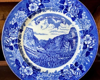 May include: A round ceramic plate featuring a blue and white transferware design. The centre depicts a landscape of Yosemite Valley, California, with a banner containing the text YOSEMITE VALLEY, CALIFORNIA. The rim has a floral pattern.