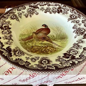 May include: A Spode porcelain serving bowl with a pheasant design. The bowl features a cream-colored interior with a detailed illustration of two pheasants in a grassy field. The rim is decorated with a brown floral pattern. The bowl is on top of a Spode box.