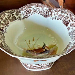 May include: A cream-colored ceramic bowl with a scalloped rim featuring a brown floral and lattice pattern. The interior base displays a painted scene of a duck near cattails. The exterior base also features a matching brown floral border.