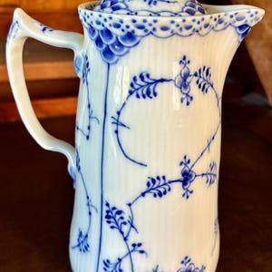May include: A white porcelain creamer with a ribbed body and a matching lid. The piece features a delicate blue floral pattern, a scalloped rim, and a curved handle. The design includes intricate blue painted vines and flowers on the exterior.