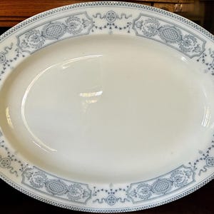 May include: An oval-shaped white ceramic serving platter featuring a decorative border with a repeating blue and white pattern. The rim displays intricate floral motifs, geometric lattice designs, and delicate swags in a soft blue color.