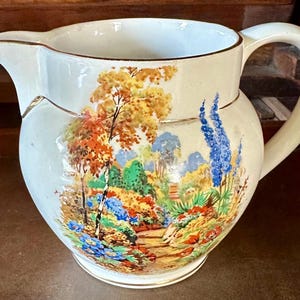 May include: Cream-colored ceramic pitcher with a landscape scene featuring trees, flowers, and a pathway. The pitcher has a rounded body, a curved handle, and a spout. Gold trim accents the rim and base.
