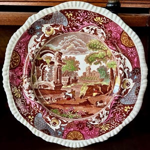 May include: A decorative ceramic plate with a ruffled edge. The plate features a landscape scene in shades of brown, pink, and green. The rim has a white, scalloped edge. The plate's design includes floral and scroll patterns.