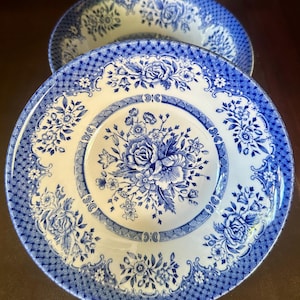 May include: Two white ceramic plates with blue floral patterns. The plates have a blue floral design in the center and around the edges. The plates are on a dark brown surface.