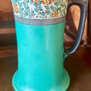 May include: A teal green ceramic pitcher with a white band and floral pattern around the top. The handle is black.