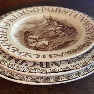 May include: Three white ceramic plates with brown trim and a brown alphabet border. The top plate features a brown illustration of two figures in a landscape.