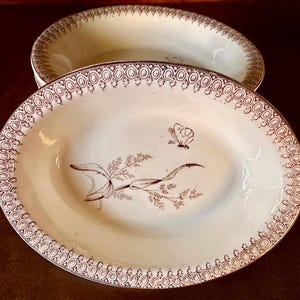 May include: Two antique oval ceramic serving dishes with a brown floral and butterfly design on a white background. The dishes have a brown scalloped edge.