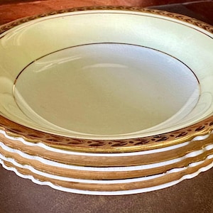 May include: A set of four white ceramic bowls with gold trim. The bowls are stacked on top of each other. The bowls have a scalloped edge and a gold design around the rim.