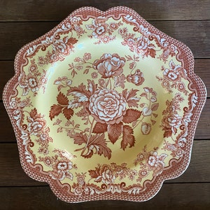 May include: A decorative, scalloped-edge serving platter with a floral design. The plate features a yellow center with a large brown rose and leaf pattern. The border is brown with a repeating floral and geometric design.