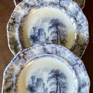 May include: Three antique blue and white plates with a castle scene. The plates have a scalloped edge and a detailed border design. The central image depicts a castle, trees, and a bridge. The plates are arranged in a vertical stack.