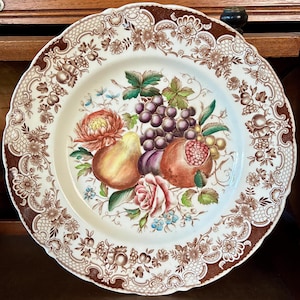 May include: A round ceramic plate with a scalloped edge featuring a brown and white floral border. The center displays a colorful illustration of a pear, a pomegranate, a cluster of purple grapes, and various flowers including a pink rose.