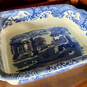 May include: A rectangular ceramic baking dish featuring a white base with a blue transferware pattern. The rim displays a dense floral and leaf motif, while the center depicts a detailed landscape scene with ruins, trees, and figures.