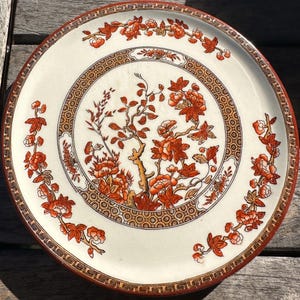 May include: A round, cream-colored plate with a detailed floral design in shades of orange and brown. The plate features a central floral motif surrounded by a geometric border and a decorative rim. The design includes stylized flowers and branches.