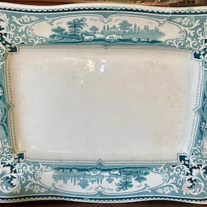 May include: A rectangular ceramic serving platter featuring a white center and a wide, ornate border in teal green. The border displays intricate scrollwork, floral motifs, and detailed landscape scenes with trees, buildings, and grazing animals.