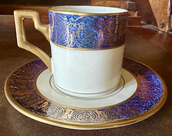 May include: A white teacup and saucer set with gold trim and a blue and gold patterned design. The cup has a square handle. The saucer and the upper part of the cup have a blue and gold pattern. The set is likely vintage.