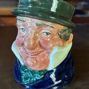 May include: A ceramic character jug depicting a person with a flushed complexion, wearing a dark green hat, a white high-collared shirt, a green necktie, and a deep blue coat. The figure has a smiling expression with visible facial features.