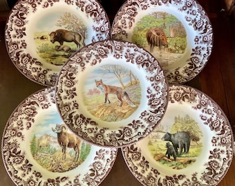 May include: A set of five white ceramic plates featuring a brown floral border pattern. Each plate displays a different colored wildlife illustration in the center, including a buffalo, a moose, a fox, a bighorn sheep, and a black bear with a cub.