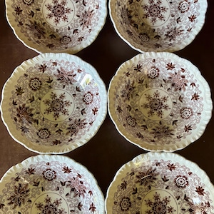 May include: A set of six round ceramic bowls featuring a scalloped edge and a cream-colored base. Each bowl is decorated with a detailed dark brown floral pattern that includes intricate blossoms, leaves, and swirling vine motifs throughout.
