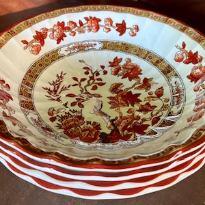 May include: A stack of four decorative ceramic bowls. The bowls have a white interior with a floral and tree design in shades of red and brown. The rims are scalloped and have a red and white striped pattern.