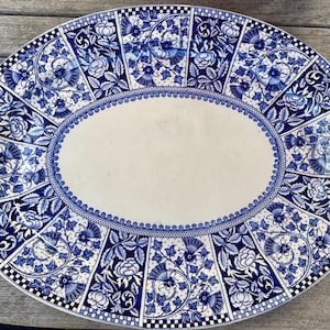 May include: Oval serving platter with a blue and white floral design. The platter features a central oval area surrounded by a detailed border of flowers, leaves, and geometric patterns. The rim has a checkered pattern.