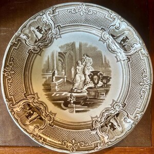 May include: A vintage decorative plate with a sepia-toned illustration of classical figures in an architectural setting. The plate features a detailed border with ornate scrollwork and geometric patterns, adding to its antique aesthetic.