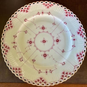 May include: A round white porcelain plate featuring a delicate red floral pattern and a central circular motif. The rim is scalloped with a pierced lattice design, accented by red scale-like patterns and a thin gold-colored edge trim.
