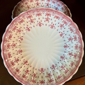 May include: Two white ceramic plates with a red floral and fleur-de-lis pattern. The plates have a scalloped edge and a textured, ridged center. The red design borders the edge and features small floral and fleur-de-lis motifs.
