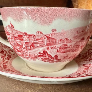 May include: A vintage teacup and saucer set with a red and white floral pattern. The cup and saucer feature a scene of a small town with houses and trees.