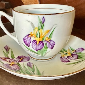May include: White teacup and saucer set with a gold rim. The cup and saucer are decorated with purple and yellow iris flowers and green leaves. A classic floral design for tea time.