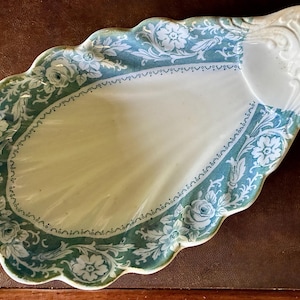 May include: A shell-shaped ceramic serving dish featuring a white center and a scalloped edge decorated with a teal and white floral pattern. The dish has a raised, embossed fan-like design at the narrow end and a smooth, glossy finish throughout.