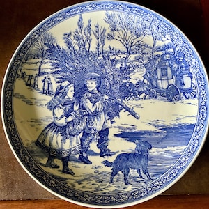 May include: A blue and white decorative plate featuring a winter scene with children carrying a Christmas tree, a dog, and a horse-drawn carriage. The plate has a detailed border design.