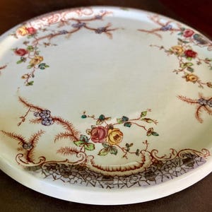 May include: A round, off-white ceramic plate with a floral design. The plate features a border of brown and black lace-like patterns and is adorned with colorful roses, leaves, and berries. The plate has a slightly raised edge.