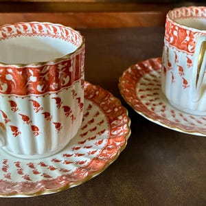 May include: Two porcelain teacups and matching saucers featuring a white base with vertical fluted ridges. The set is decorated with a repeating orange-red floral pattern and a solid orange-red band at the top, finished with gold-trimmed scalloped edges.