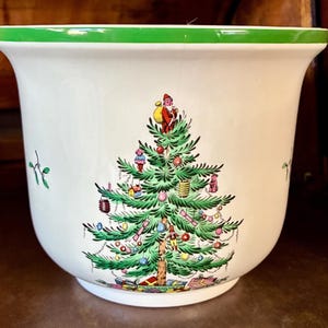 May include: A white ceramic planter with a green rim featuring a central illustration of a decorated Christmas tree topped with Santa Claus and surrounded by colorful ornaments, small figures, and wrapped gifts at the base of the tree.