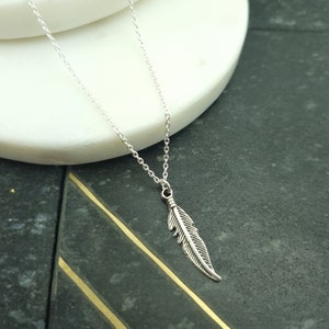 May include: A silver chain necklace with a delicate feather pendant. The necklace is on a white surface with a black background.