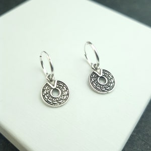 May include: A pair of silver hoop earrings with a small, round, silver charm hanging from each hoop. The charms have a detailed, intricate design.