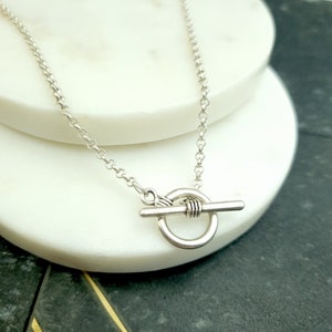 May include: A silver chain necklace with a toggle clasp. The clasp is a round ring with a bar that slides through it.