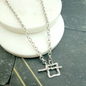 May include: A silver chain necklace with a silver toggle clasp pendant. The pendant is a simple square shape with a textured surface.