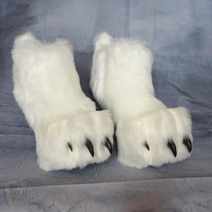 Puede incluir: Un par de zapatillas blancas y peludas con forma de pata de oso. Cada zapatilla tiene una parte superior redondeada y un dise&ntilde;o de pata con garras negras. Las zapatillas est&aacute;n hechas de un material suave y afelpado.