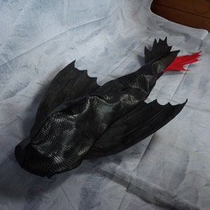 May include: A black dragon plush toy featuring a textured scale pattern on the body, large wings, and a distinct red tail fin. The creature is depicted in a flying position with its wings spread wide, showcasing the detailed black fabric construction.