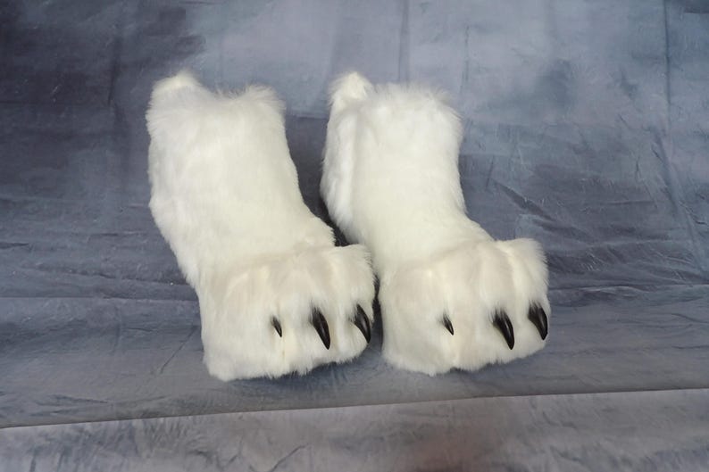 Puede incluir: Un par de zapatillas blancas y peludas con forma de pata de oso. Cada zapatilla tiene una parte superior redondeada y un dise&ntilde;o de pata con garras negras. Las zapatillas est&aacute;n hechas de un material suave y afelpado.
