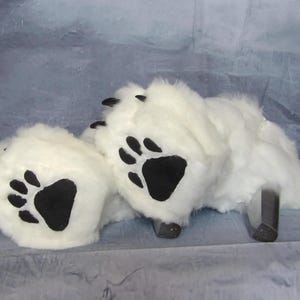 Puede incluir: Zapatillas blancas de imitaci&oacute;n de piel de oso con almohadillas y garras negras. Las zapatillas tienen una textura esponjosa y est&aacute;n dise&ntilde;adas para parecerse a las patas de un oso. El fondo es de color azul gris&aacute;ceo claro.