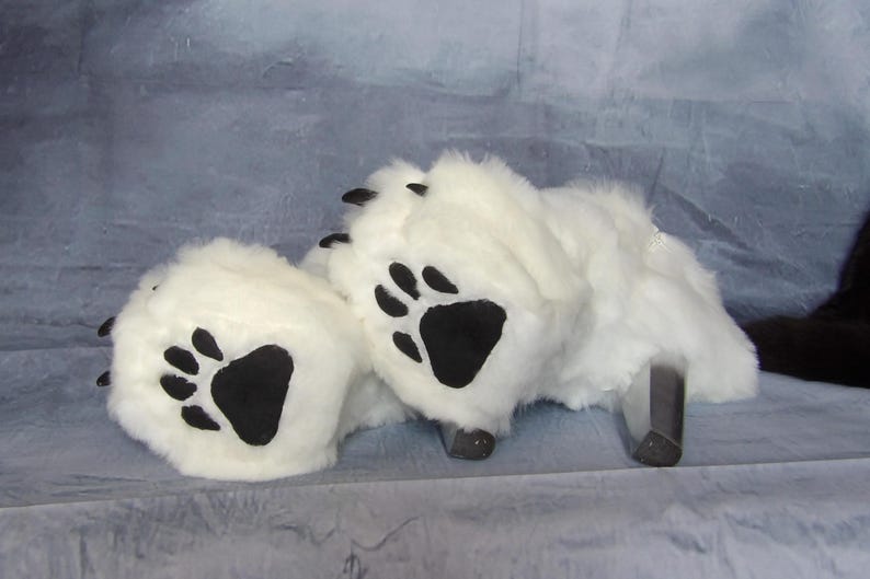 Puede incluir: Zapatillas blancas de imitaci&oacute;n de piel de oso con almohadillas y garras negras. Las zapatillas tienen una textura esponjosa y est&aacute;n dise&ntilde;adas para parecerse a las patas de un oso. El fondo es de color azul gris&aacute;ceo claro.