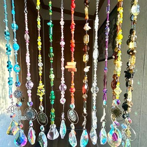 May include: A collection of beaded curtains in various colors, including blue, pink, green, red, and yellow. Each strand features different shapes and sizes of beads, with crystal pendants and decorative charms at the bottom. The curtains hang in front of a wooden door.