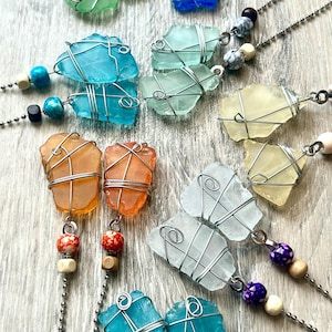 May include: A collection of handmade necklaces featuring sea glass in various colors, including blue, green, orange, and yellow. Each piece of sea glass is wrapped in silver wire and strung on a chain with decorative beads. The necklaces are displayed on a light wood surface.