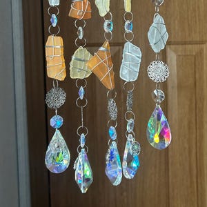 May include: A decorative hanging mobile featuring sea glass pieces in shades of orange, yellow, and white, wrapped in silver wire. The mobile also includes iridescent crystal teardrops and silver filigree accents, creating a shimmering effect.