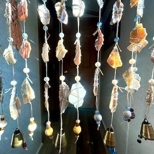 May include: A hanging mobile featuring multiple vertical strands of wire-wrapped stones, beads, and small brass bells. The stones vary in shades of white, tan, and orange, interspersed with clear, blue, and striped beads on light-colored string.